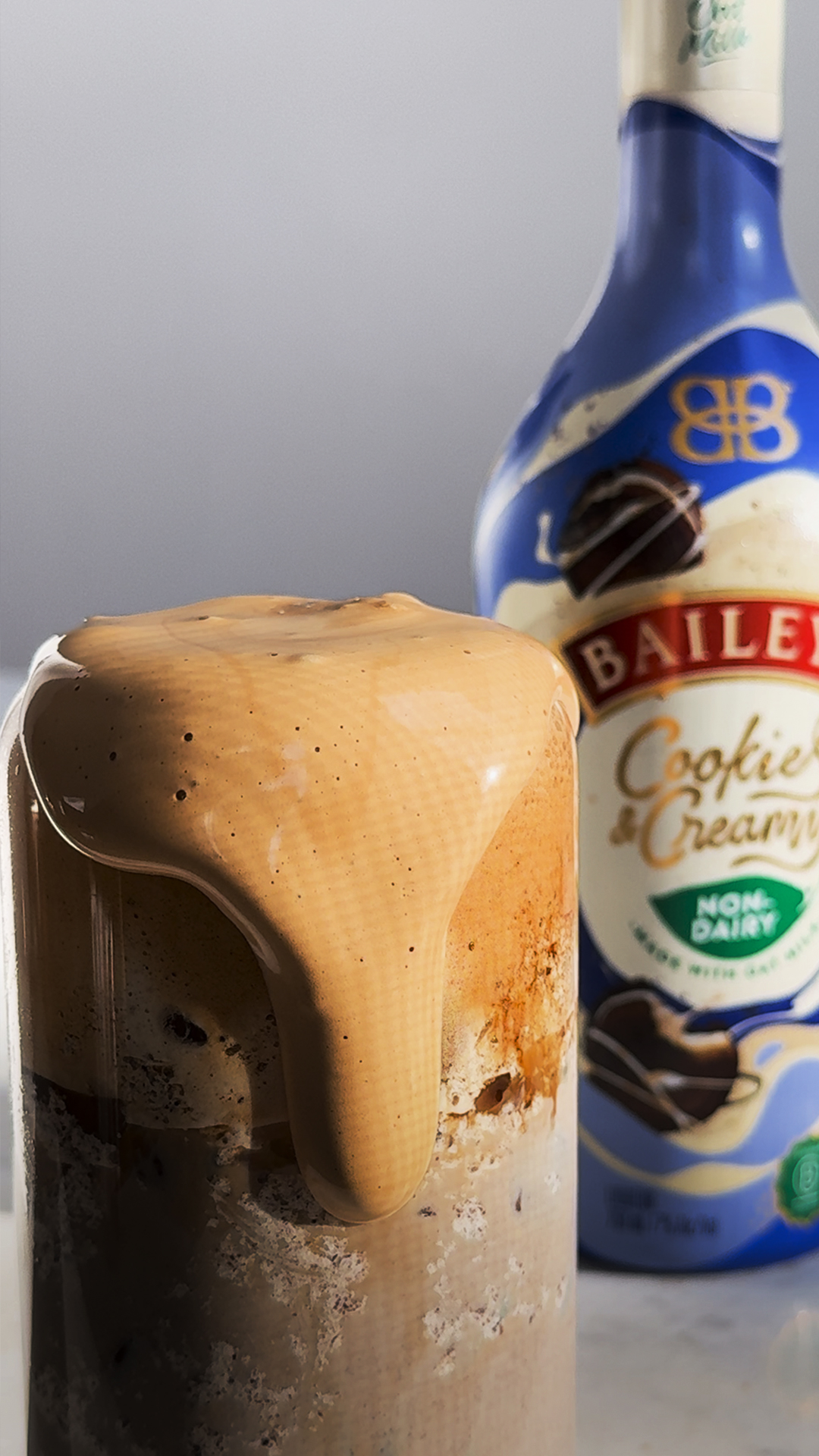 A glass filled with iced coffee topped with thick foam overflowing down the side, with a bottle of Baileys Cookie & Cream non-dairy liqueur in the background.