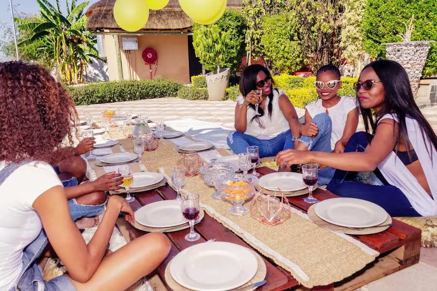 A group of women are sitting around a low outdoor table set for a meal, talking and smiling in a sunny garden setting with yellow balloons overhead.