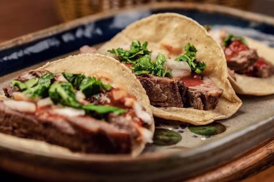 Three beef tacos topped with chopped onions, cilantro, and salsa are arranged on a ceramic plate.