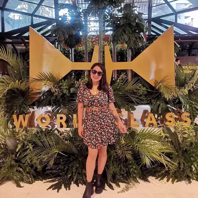 En la imagen se ve a Esteffany delante del logo del World Class, rodeado de plantas.