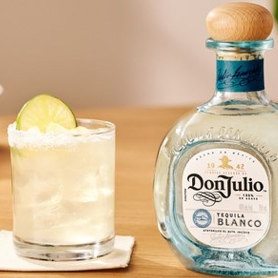 La imagen muestra una botella de Tequila Don Julio Blanco junto a vasos rellenos con el cóctel Margarita y diferentes platos de comida.