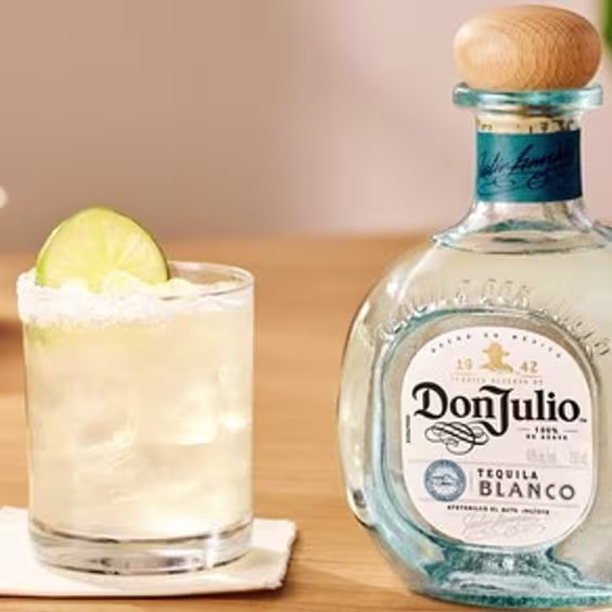La imagen muestra una botella de Tequila Don Julio Blanco junto a vasos rellenos con el cóctel Margarita y diferentes platos de comida.