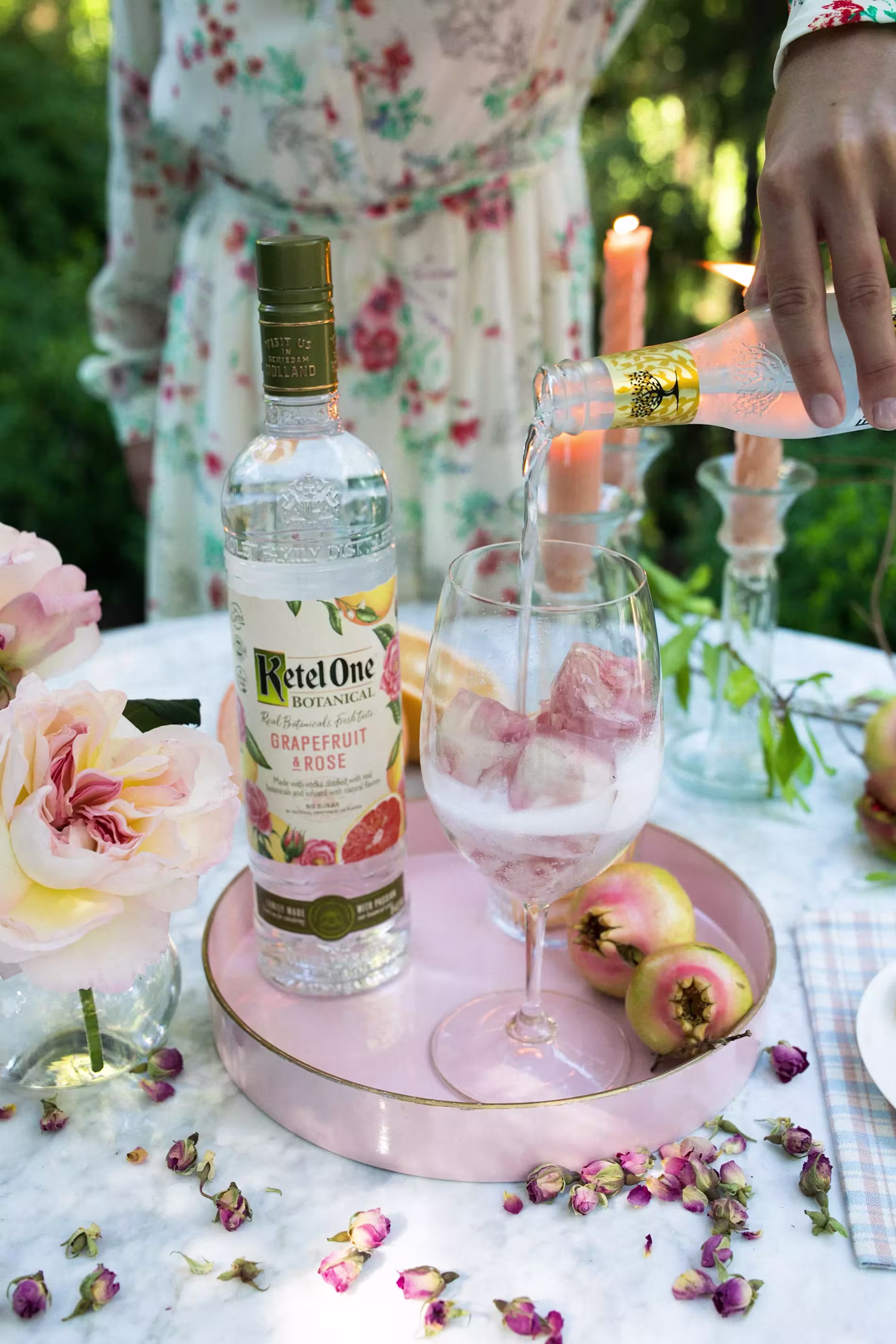 A hand pours sparkling water into a wine glass with ice next to a bottle of Ketel One Botanical Grapefruit & Rose vodka on a table decorated with flowers and fruit.