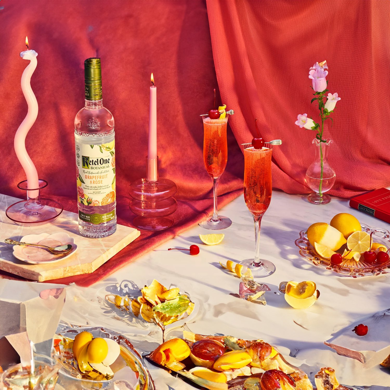A bottle of Ketel One Botanical Grapefruit & Rose vodka sits on a table with cocktails, sliced fruit, candles, flowers in vases, and broken glass on a pink and red draped backdrop.