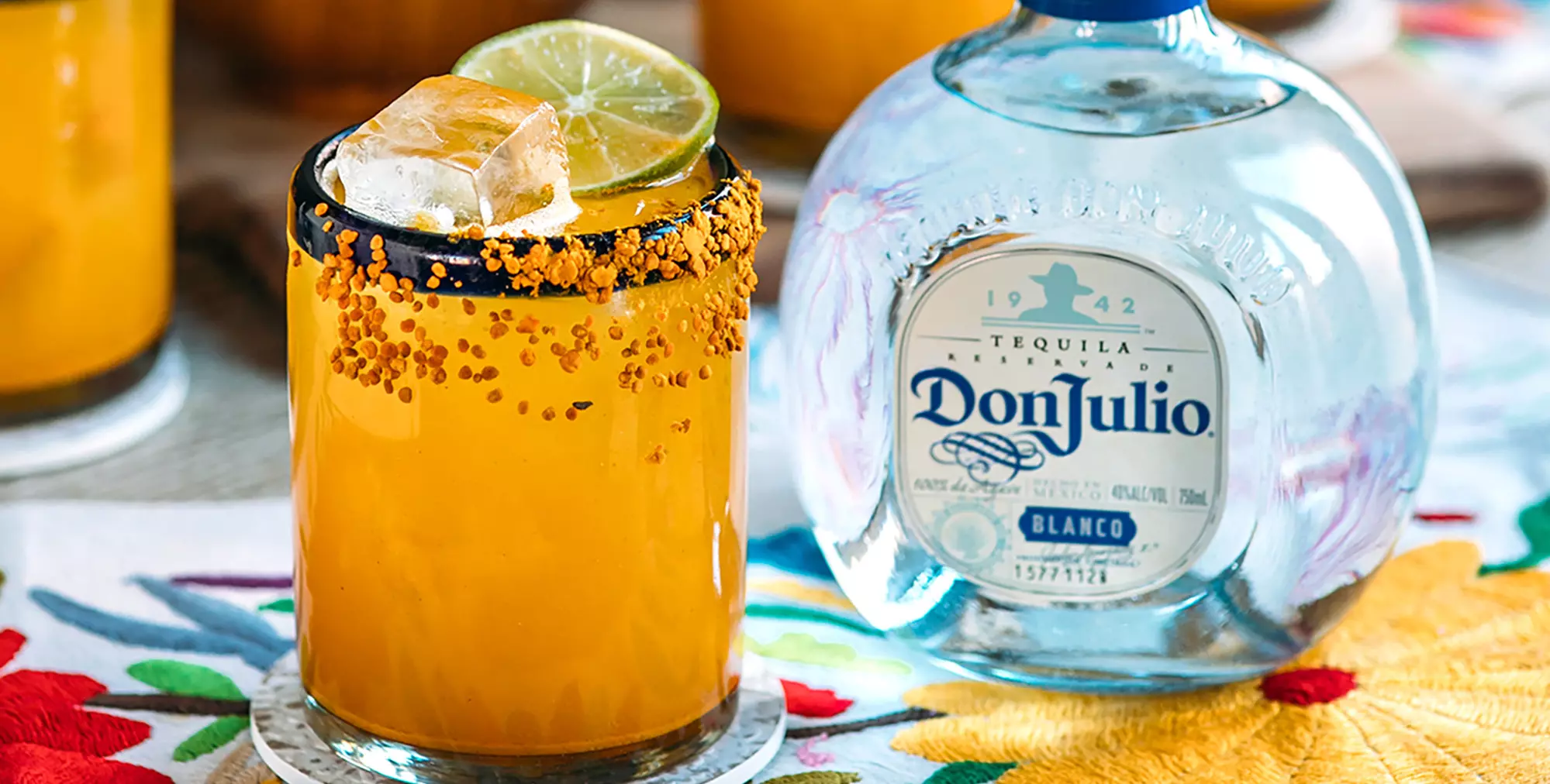 A glass of orange cocktail with ice, lime slice, and chili powder rim sits next to a bottle of Don Julio Blanco tequila on a floral tablecloth.