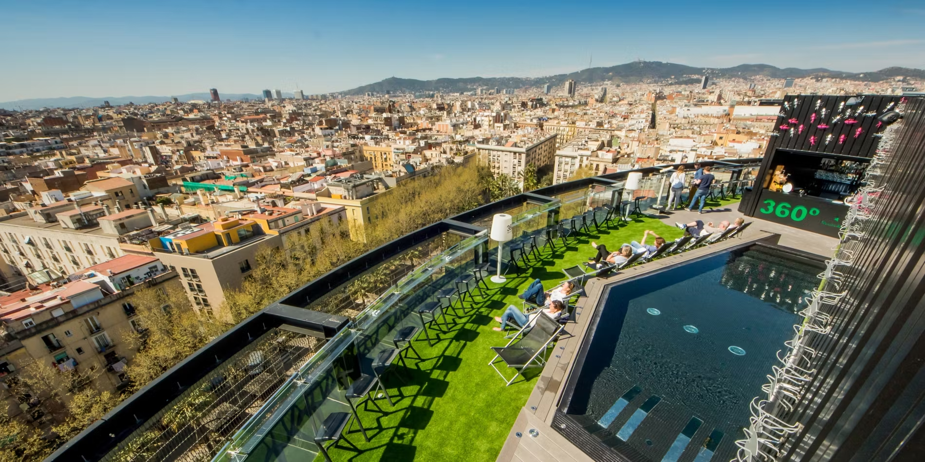 a foto muestra un espacio exterior de la 360 Barceló Raval. El área está adornada con muebles modernos, incluyendo mesas y sillas, y cuenta con una piscina