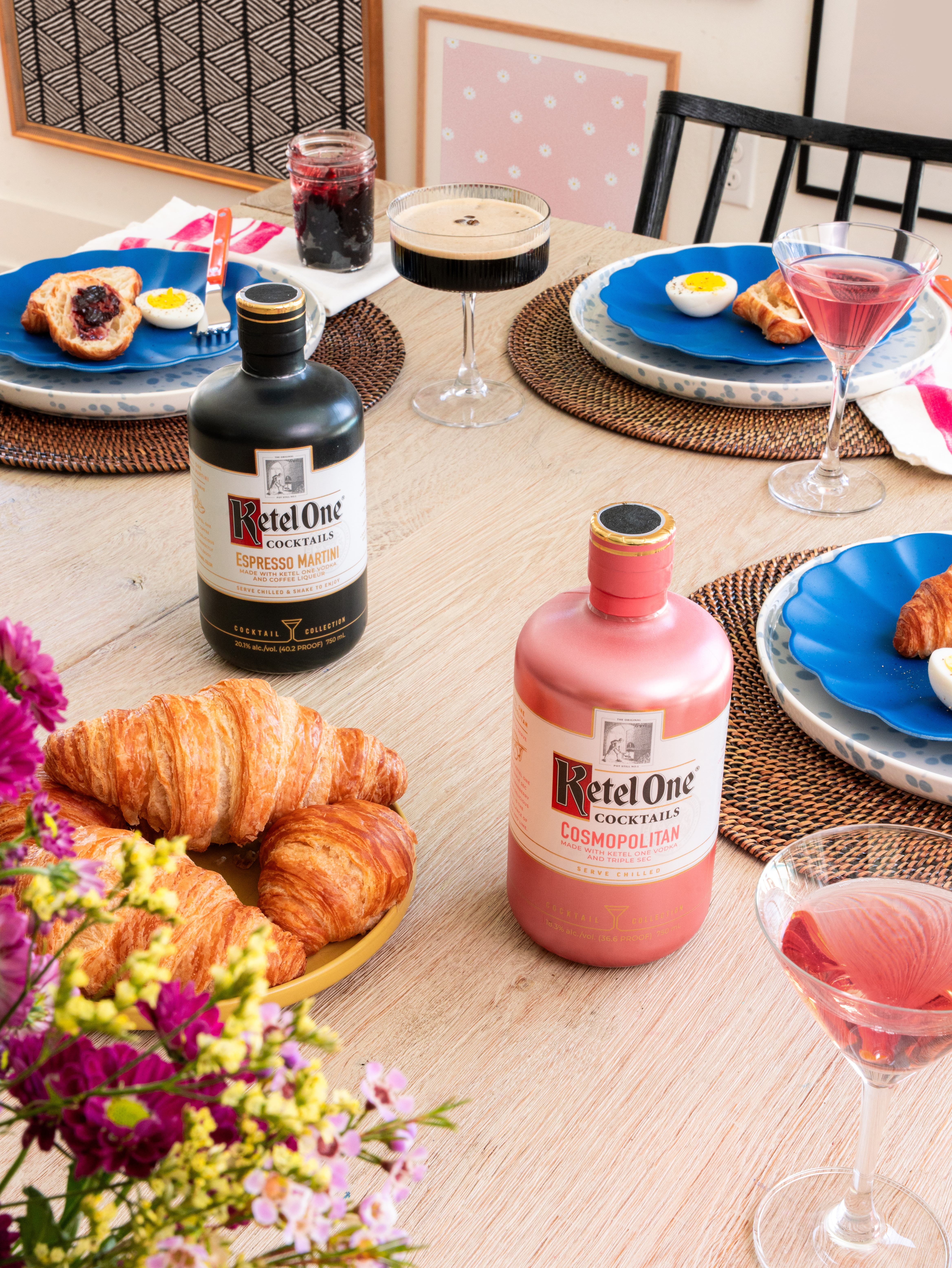 A brunch table set with croissants, pastries, blue plates, cocktails, and bottles of Ketel One Espresso Martini and Cosmopolitan. Flowers are in the foreground.