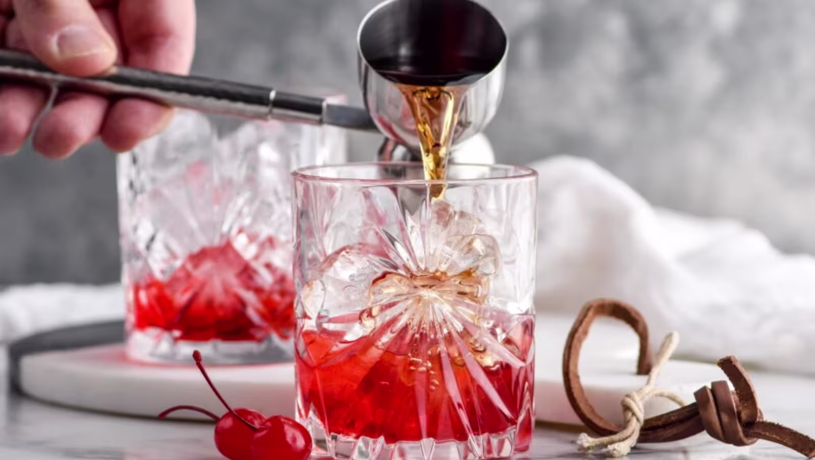 A hand pours liquor into a glass with ice and red liquid, garnished with maraschino cherries on the side. Another similar glass is in the background.