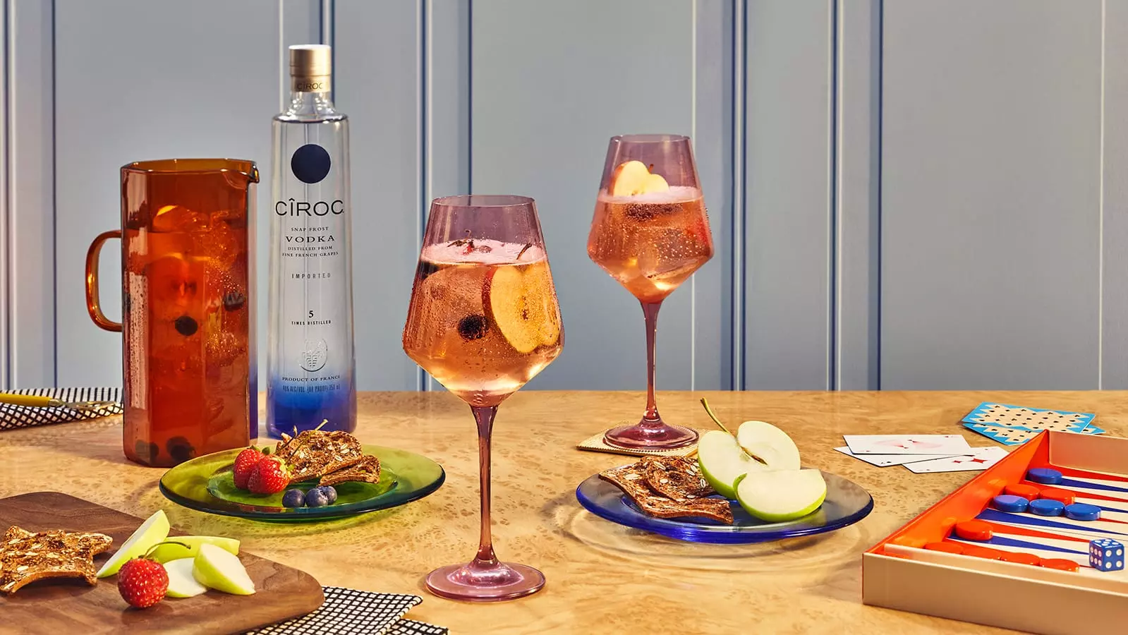 A bottle of Ciroc vodka, two cocktails with fruit, a pitcher, plates of fruit and snacks, and a backgammon game on a wooden table.
