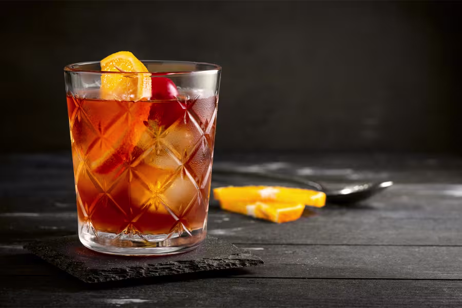 A glass of iced cocktail garnished with an orange slice and cherry sits on a dark coaster, with orange wedges and a spoon in the background.