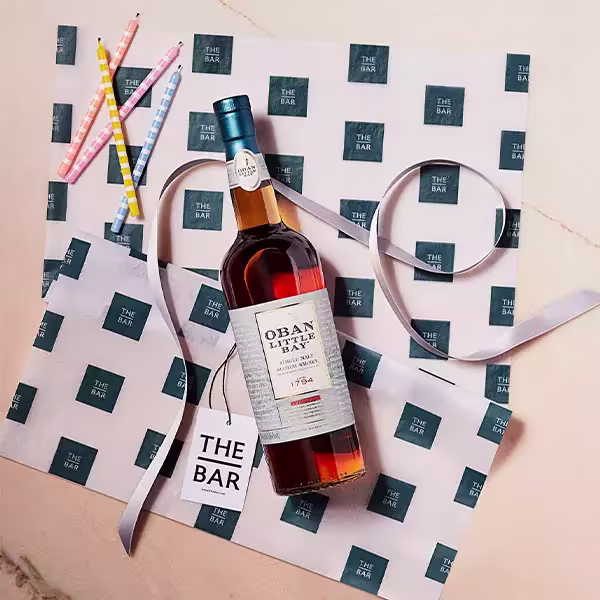 A bottle of Oban Little Bay Scotch whisky on branded wrapping paper, with a ribbon, gift tag, and colorful straws nearby.