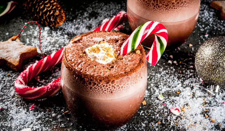 A glass of hot chocolate topped with whipped cream, garnished with a crushed cookie and a red-and-green striped candy cane, surrounded by festive holiday decorations.