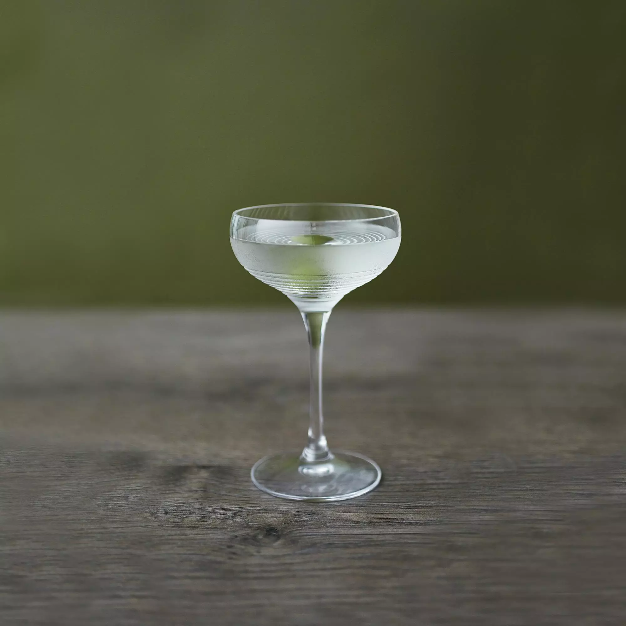 A coupe glass filled with a clear cocktail, garnished with a lemon twist, sits on a wooden surface against a green background.