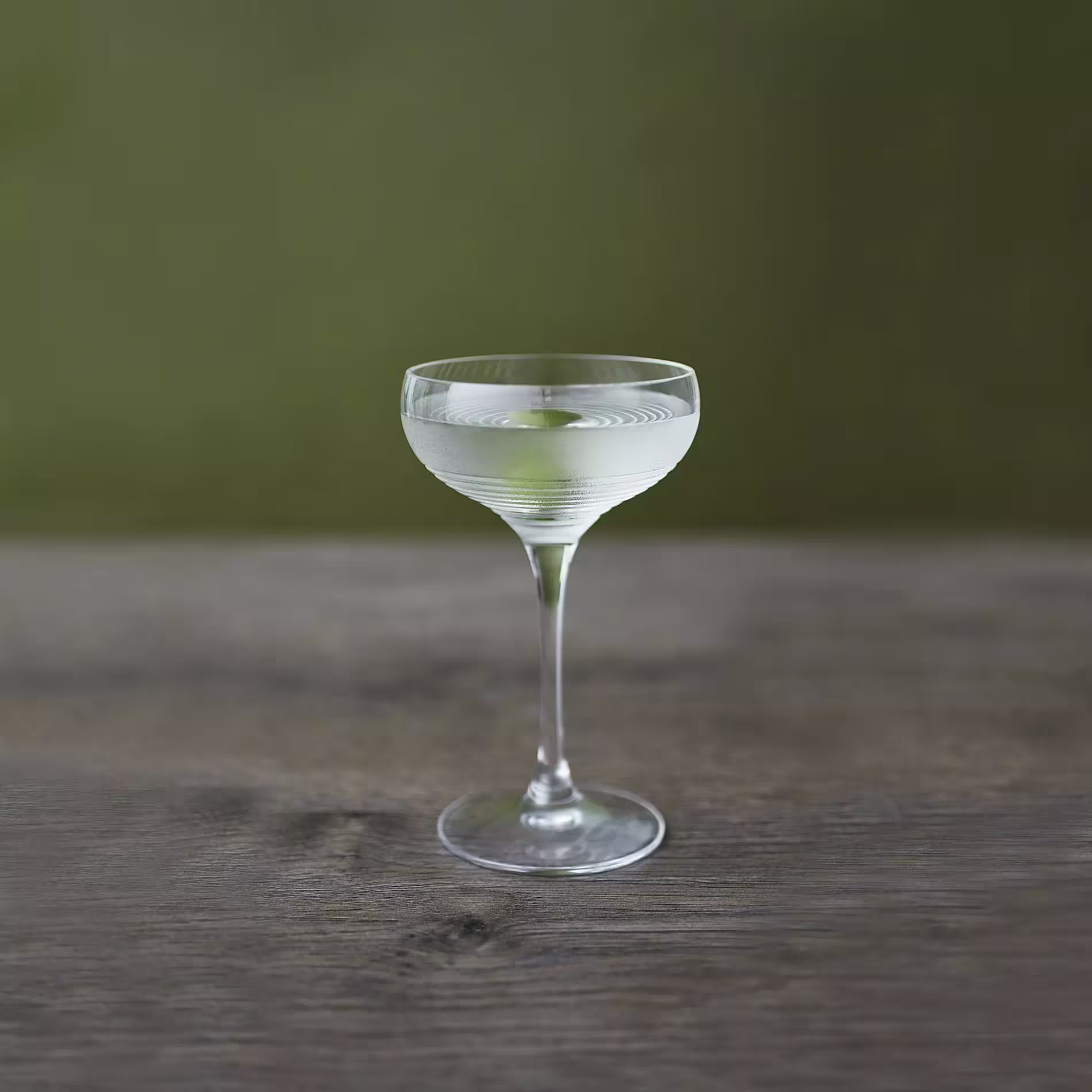 A coupe glass filled with a clear cocktail, garnished with a lemon twist, sits on a wooden surface against a green background.