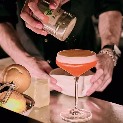 La imagen muestra un cocktail en copas de martini clásicas, con una cobertura cremosa Las bebidas están sostenidas por un soporte transparente y reflectante que da la impresión de que las copas están flotando.