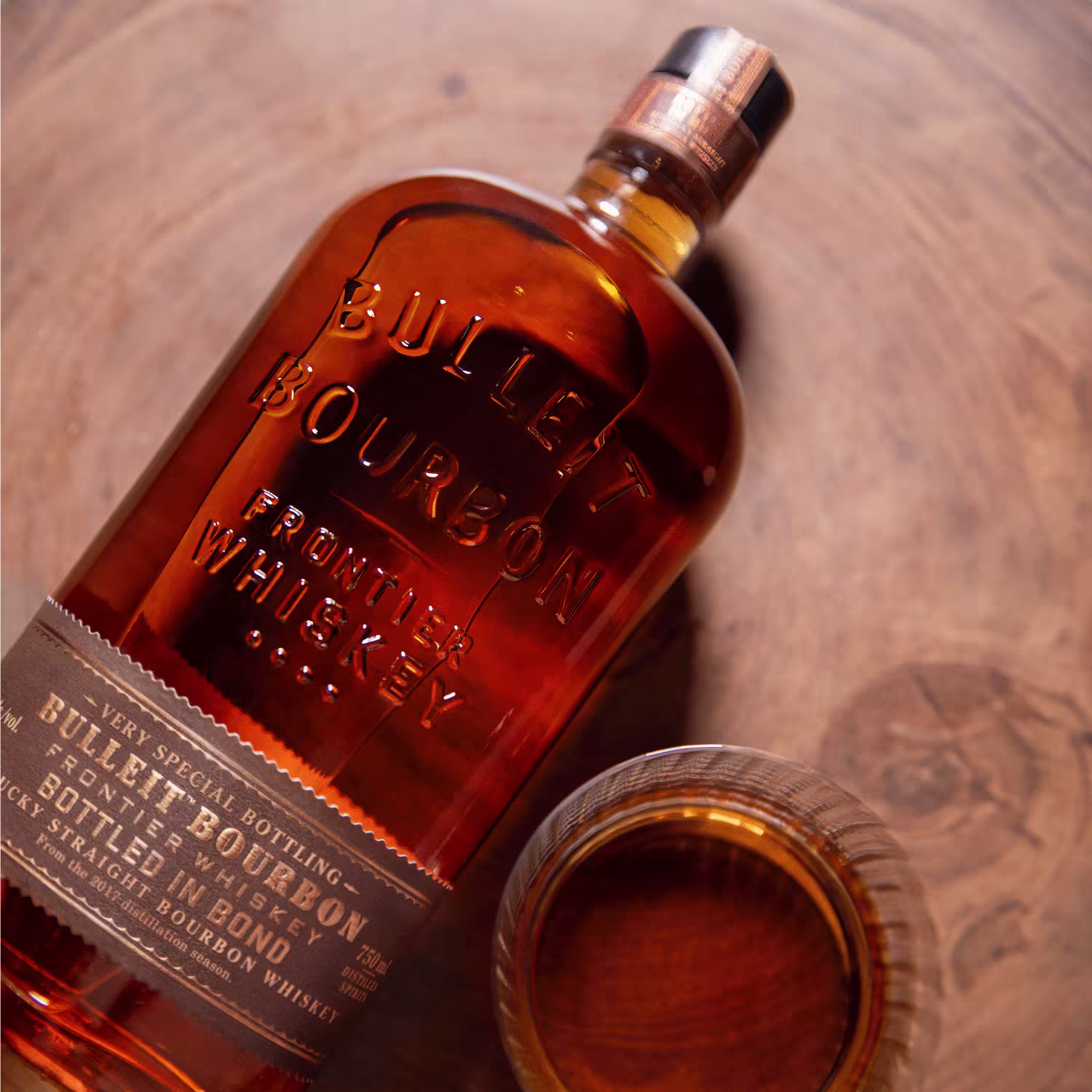 A bottle of Bulleit Bourbon Frontier Whiskey lies on its side next to a glass filled with bourbon, placed on a wooden surface.