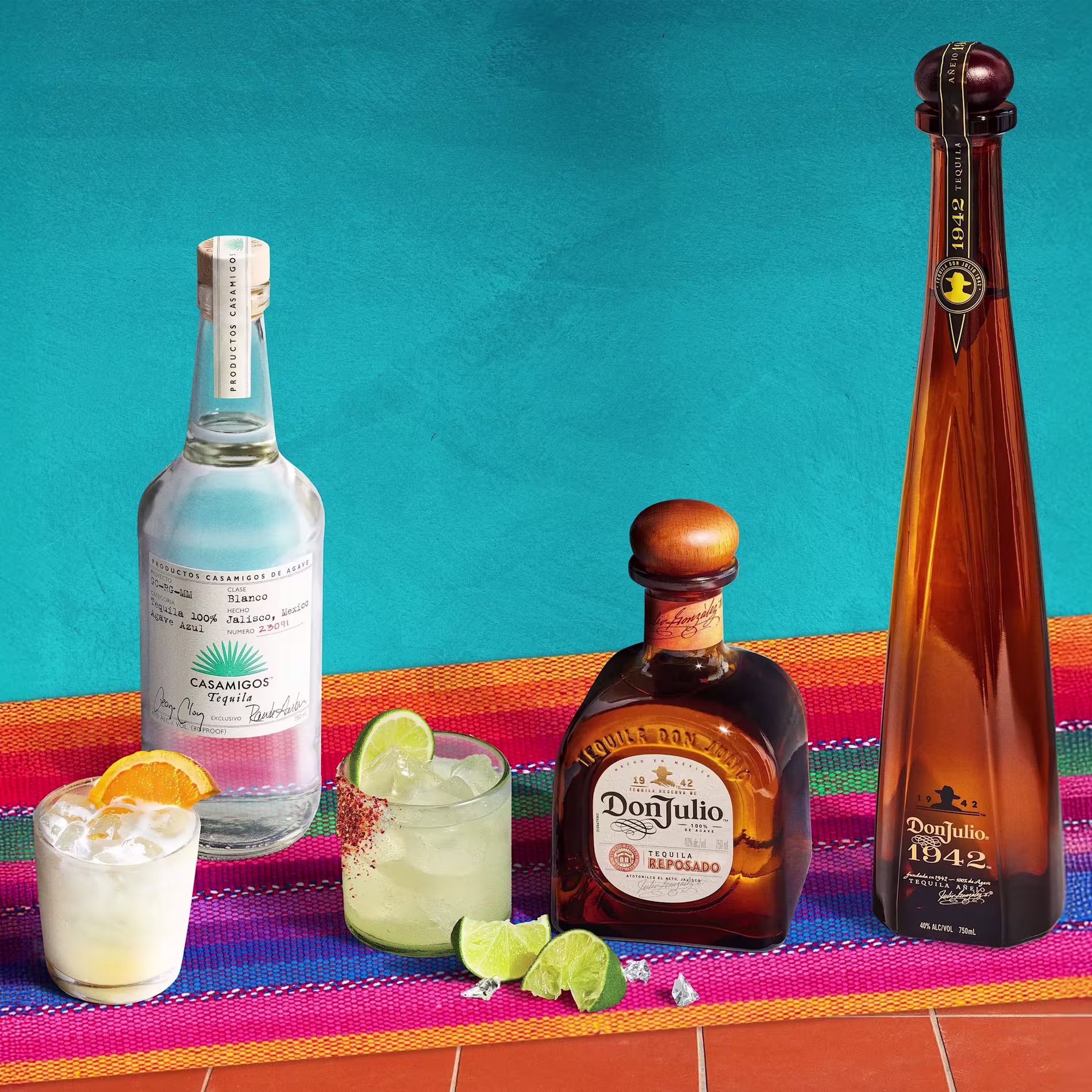 Tres botellas de tequila -Casamigos, Don Julio Reposado y Don Julio 1942- se exhiben con cócteles y gajos de lima sobre una colorida alfombra tejida con fondo azul.