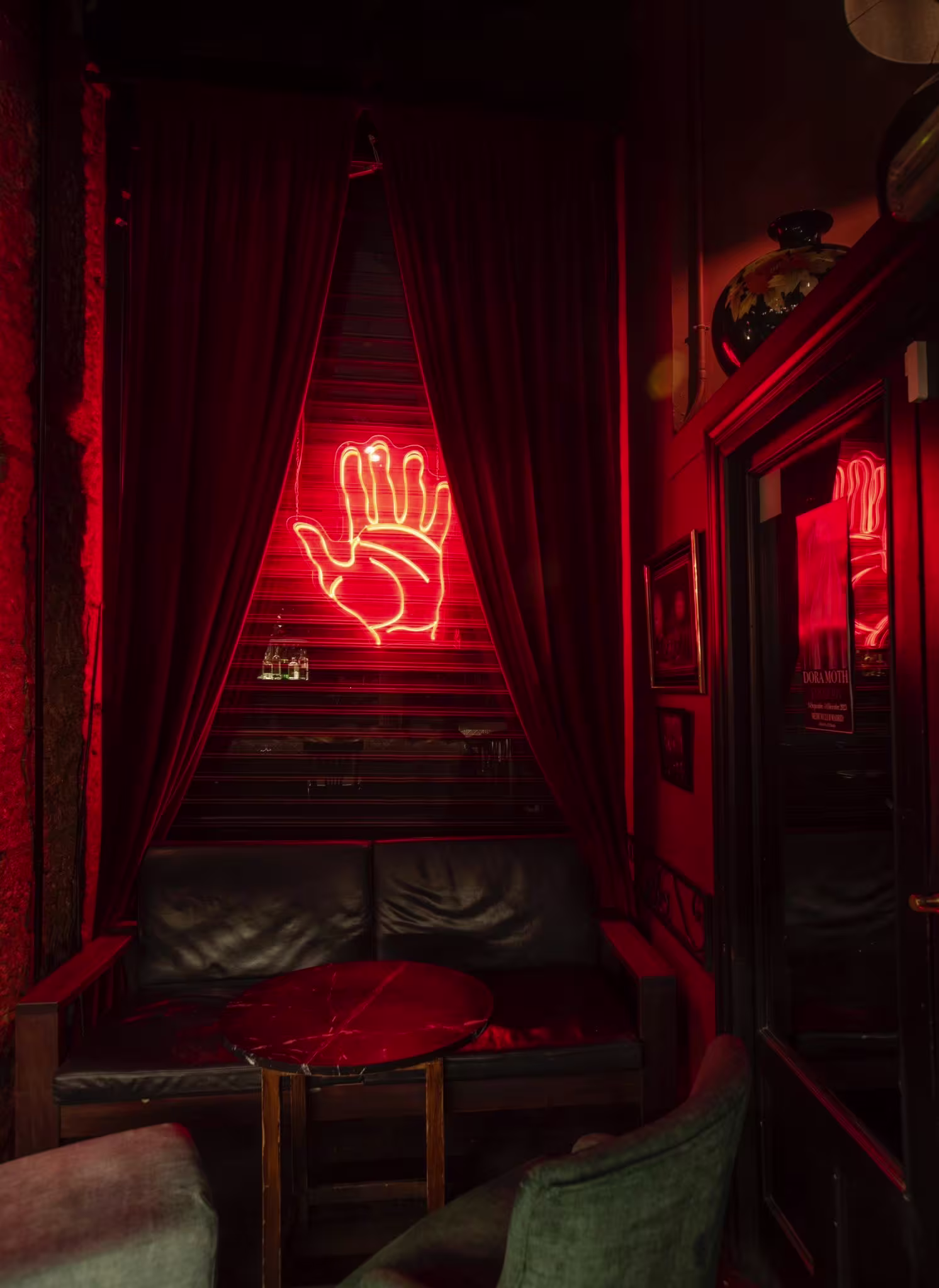 La imagen muestra el interior del Medium  CLub . Hay mesas y sillas de madera, paredes verdes con cuadros enmarcados, papel tapiz con patrones y lámparas colgantes que proporcionan una iluminación suave. 