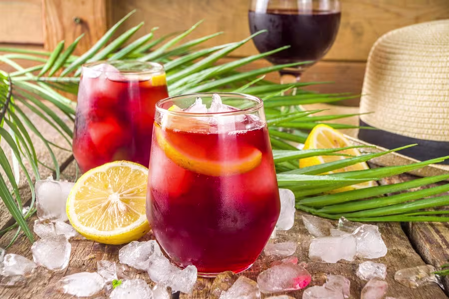 Two glasses of iced red beverage with lemon slices, surrounded by ice cubes, a lemon half, palm leaves, and a hat on a rustic wooden surface.