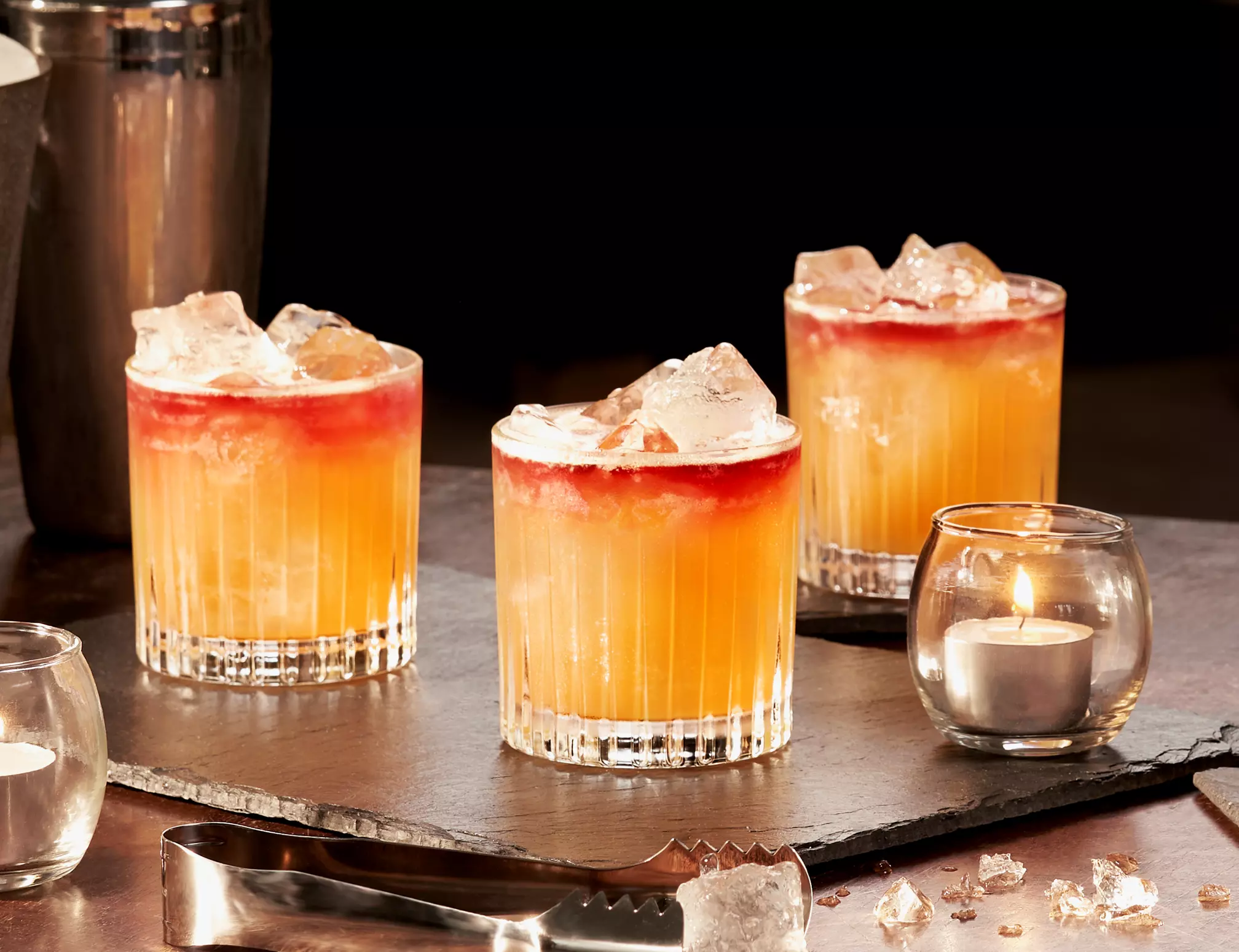 Three glasses of orange and red cocktails with ice sit on a slate tray next to a lit tealight candle, a shaker, and metal tongs on a dark surface.