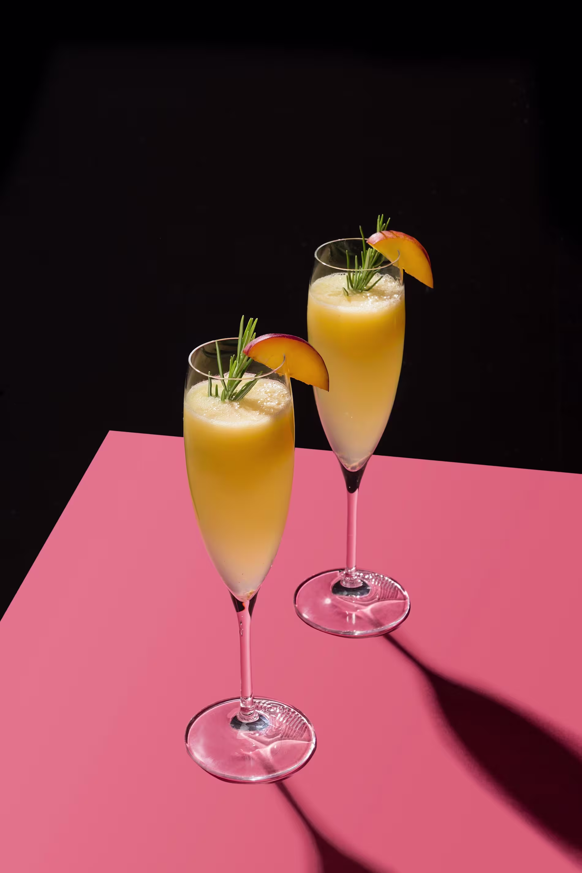 Two champagne flutes filled with a yellow-orange drink, garnished with a peach slice and rosemary sprig, on a pink surface against a black background.