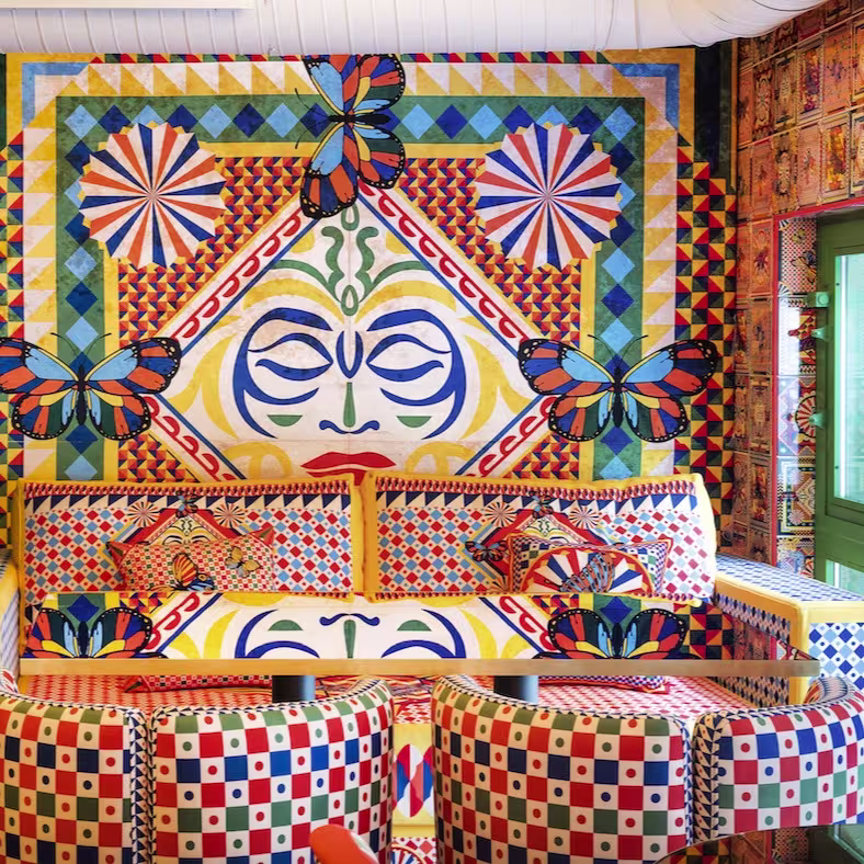 La foto muestra el espacio interior del Boom Boom Ciao, decorada con un mural colorido y simétrico.. Hay bancos con cojines a juego frente al mural y el suelo tiene baldosas con patrones. 