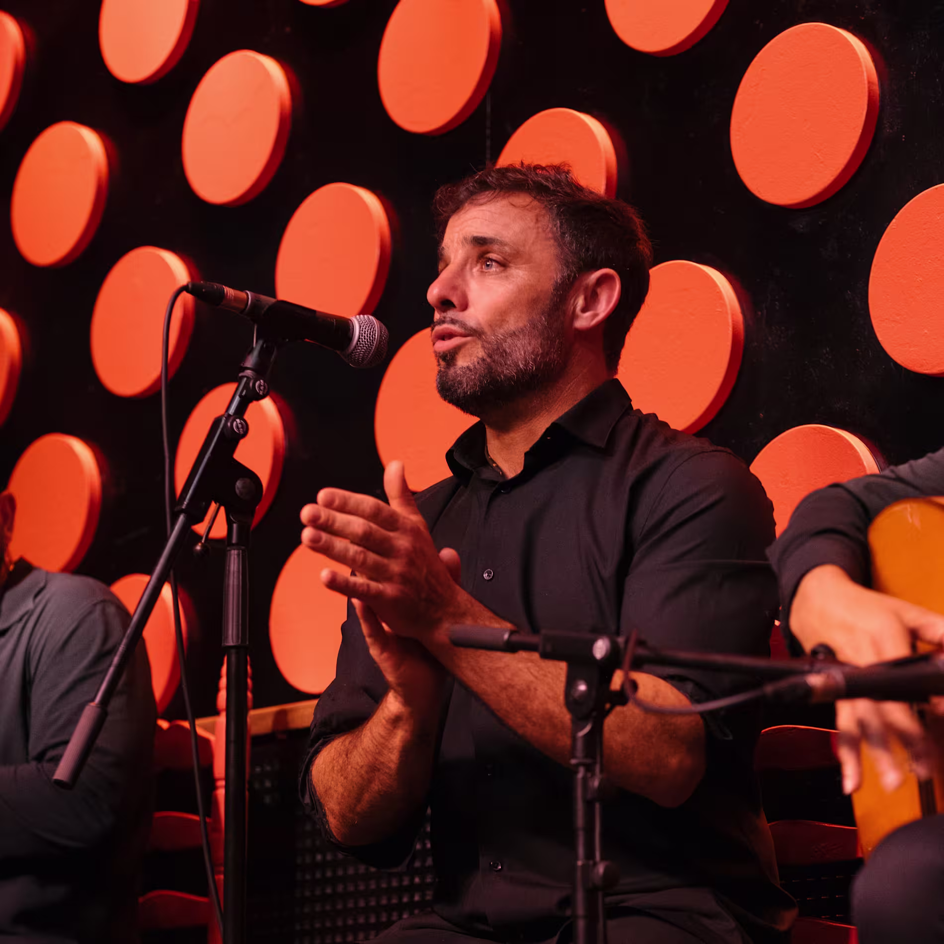 Un cantaor de flamenco aplaude con pasión frente al micrófono, rodeado de un fondo negro con grandes círculos naranjas y sillas a juego. La esencia de Tarantos en plena acción.