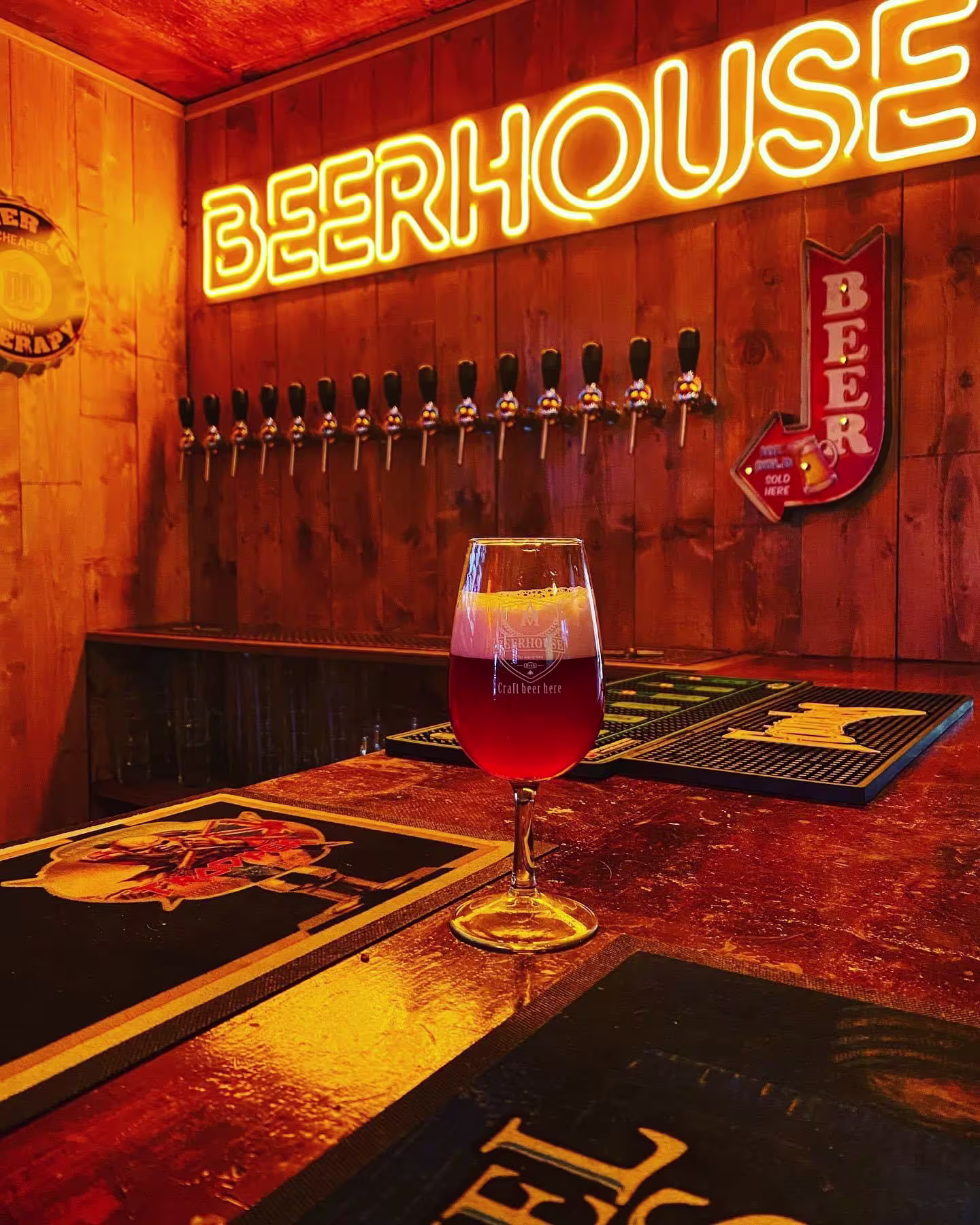La foto muestra el interior de un bar, centrado en un vaso de cerveza colocado sobre una barra de madera. Encima de la barra, hay una pantalla montada en la pared con varios grifos de cerveza, etiquetados como “BEERHOUSE”.