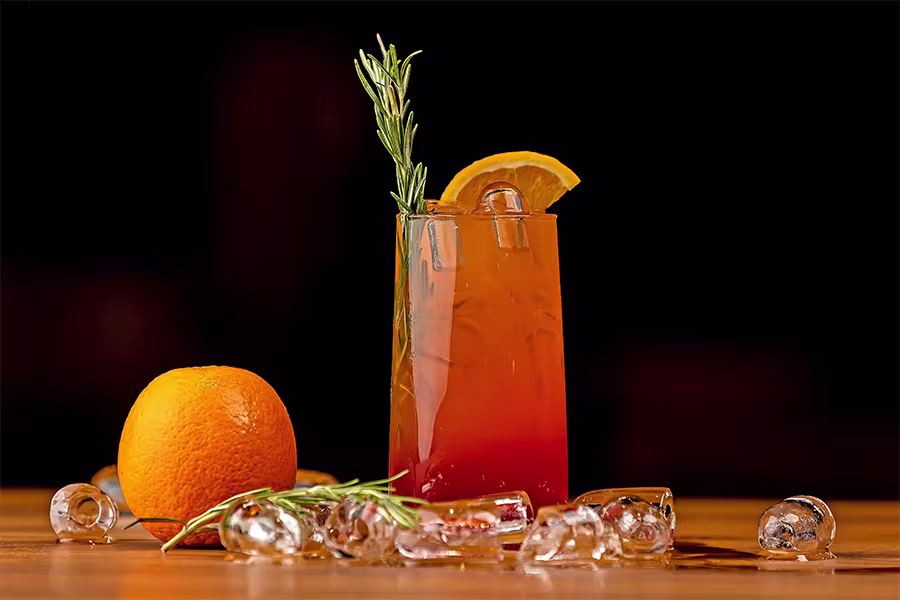 A tall glass of orange-red cocktail with ice, garnished with a rosemary sprig and orange slice, sits beside a whole orange and scattered ice cubes on a wooden surface.