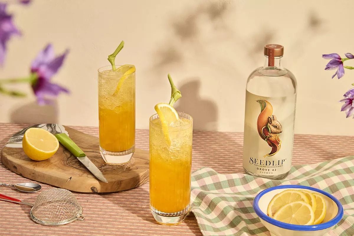 Two tall glasses with orange cocktails and lemon slices sit on a checkered cloth next to a Seedlip Grove 42 bottle, a bowl of lemon slices, a strainer, and a halved lemon on a cutting board.