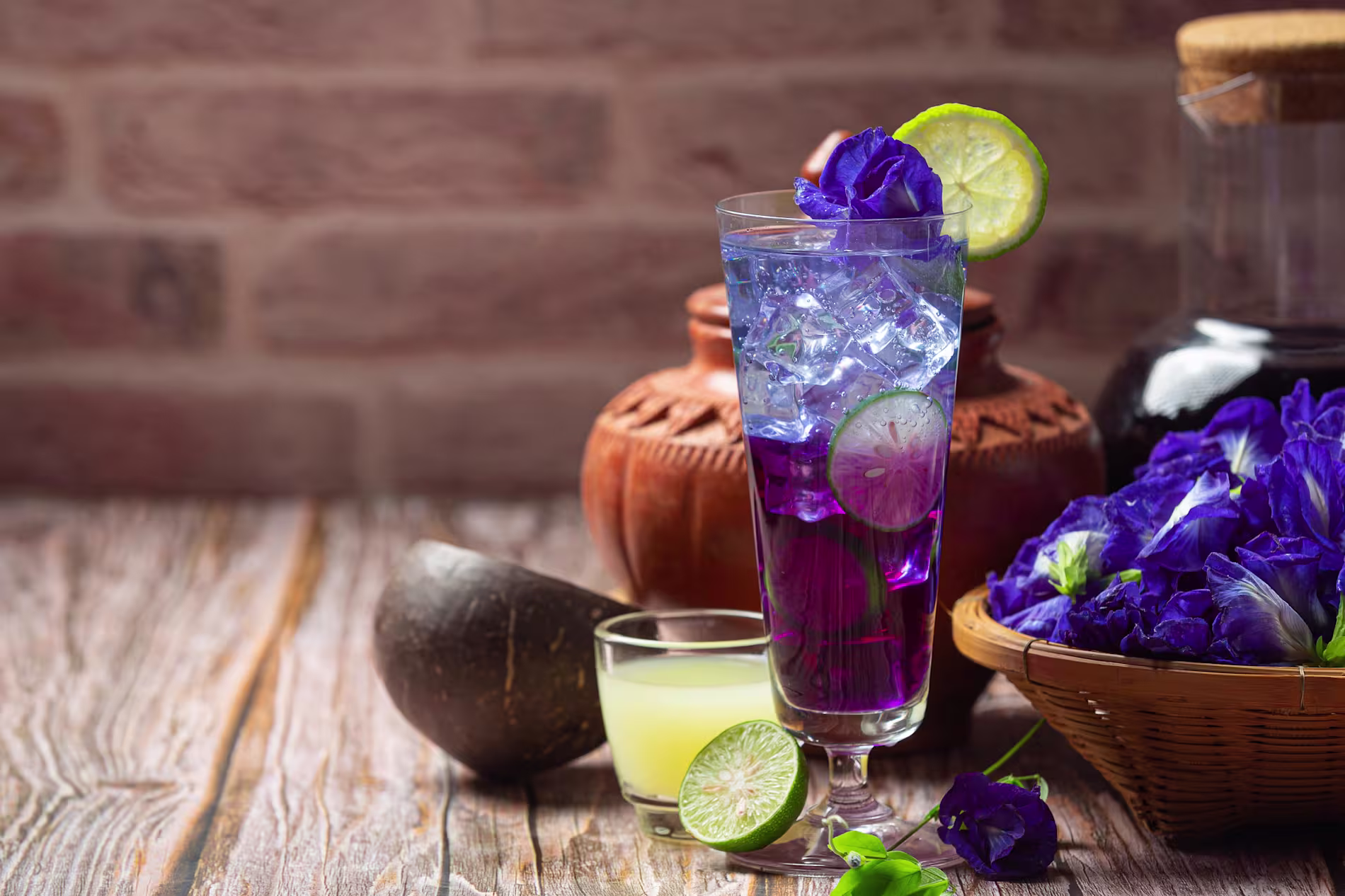 A tall glass of blue butterfly pea flower drink with ice, lime slices, and flowers sits on a rustic wooden table beside a bowl of blue flowers and a small glass of lemon juice.
