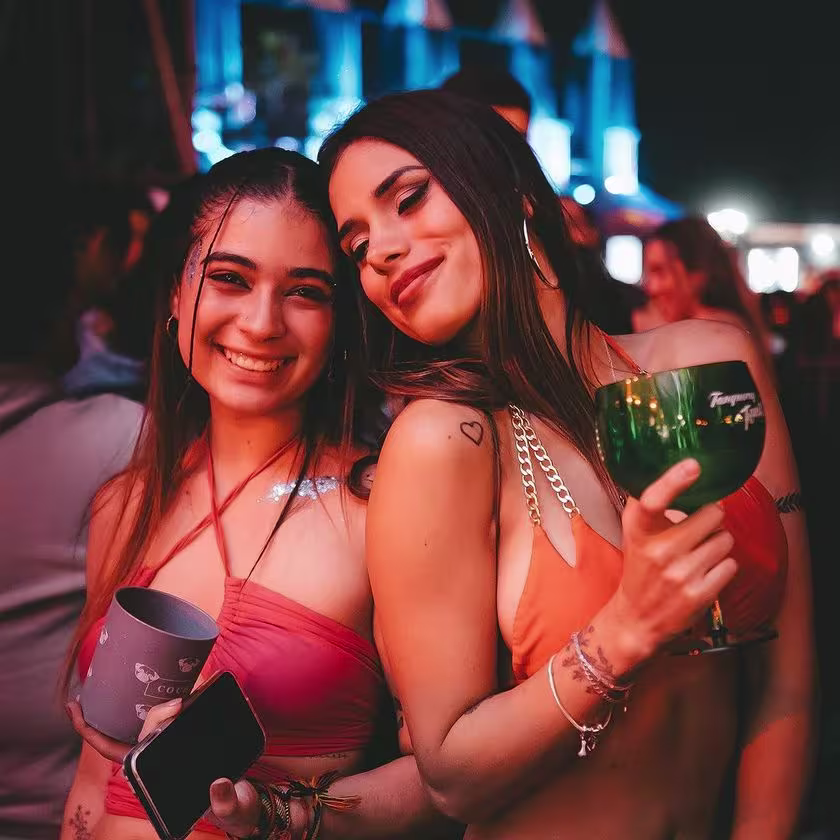 La imagen retrata a dos jóvenes sonrientes: la joven de la izquierda sostiene un vaso gris con parte de la palabra "Cochinola" escrita en él, mientras que la joven de la derecha sostiene una copa verde de Gin Tanqueray.