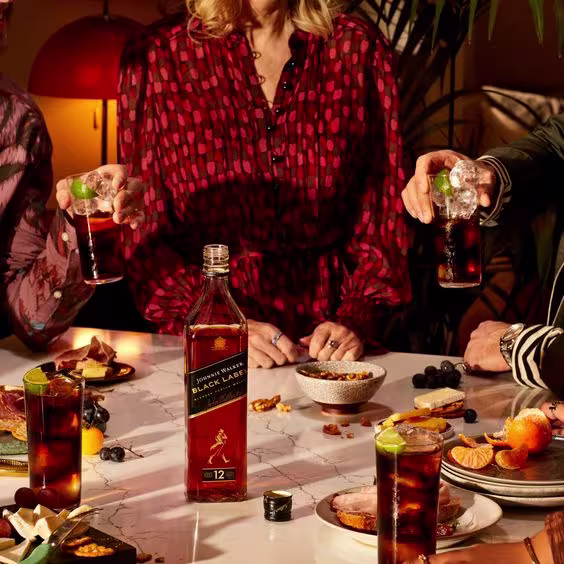 La imagen presenta una mesa con una variedad de platos, destacando en el centro una botella de whisky Johnnie Walker Black Label. Alrededor de la mesa, se observan personas, algunas de ellas sosteniendo vasos con cócteles.