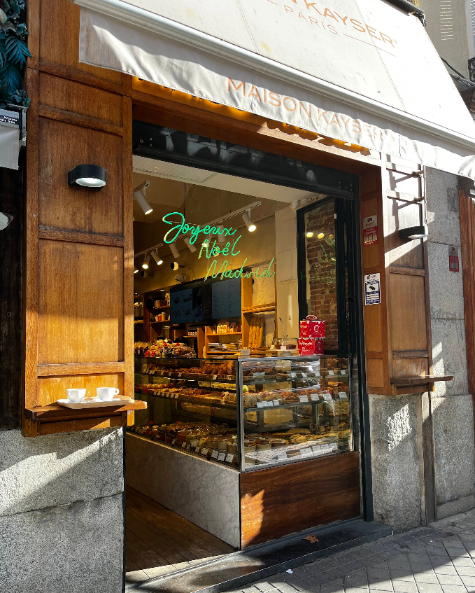 Escaparate repleto de pasteles en Maison Kayser Fernando VI, con tazas en la calle y un letrero que celebra la Navidad en Madrid.