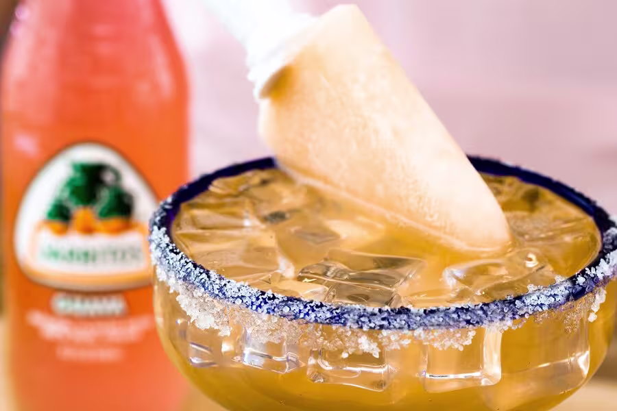 A popsicle is placed in a glass of iced beverage with a salted rim, with a bottle of Jarritos soda in the background.