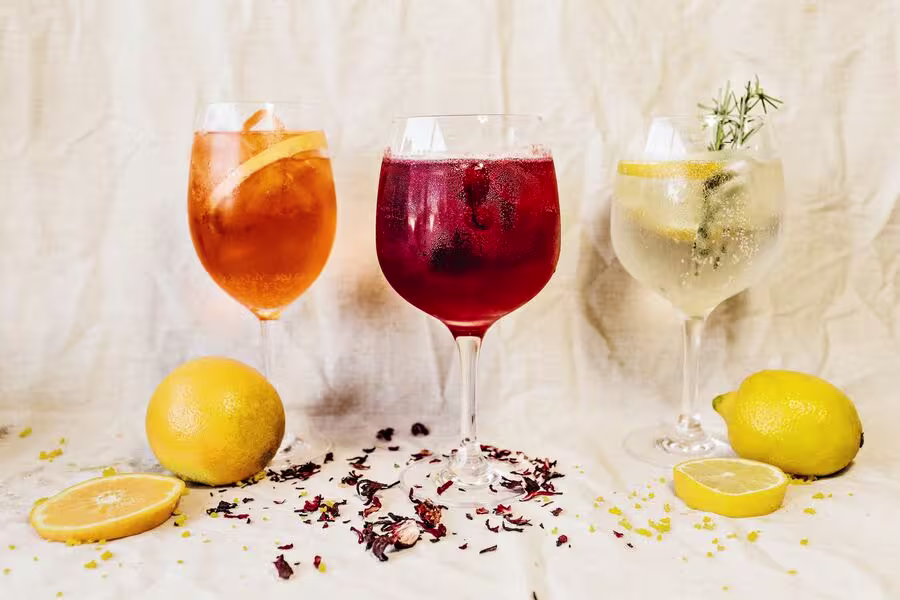 Three glasses of colorful cocktails sit on a light fabric background, surrounded by lemons, lemon halves, and scattered flower petals.