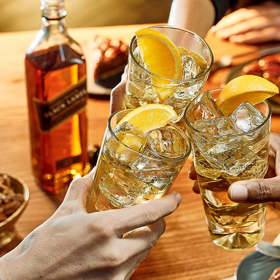 La imagen muestra tres manos sosteniendo cada una un vaso highball con whisky y limón, brindando. En segundo plano, se aprecia de manera difuminada una botella de whisky Johnnie Walker.