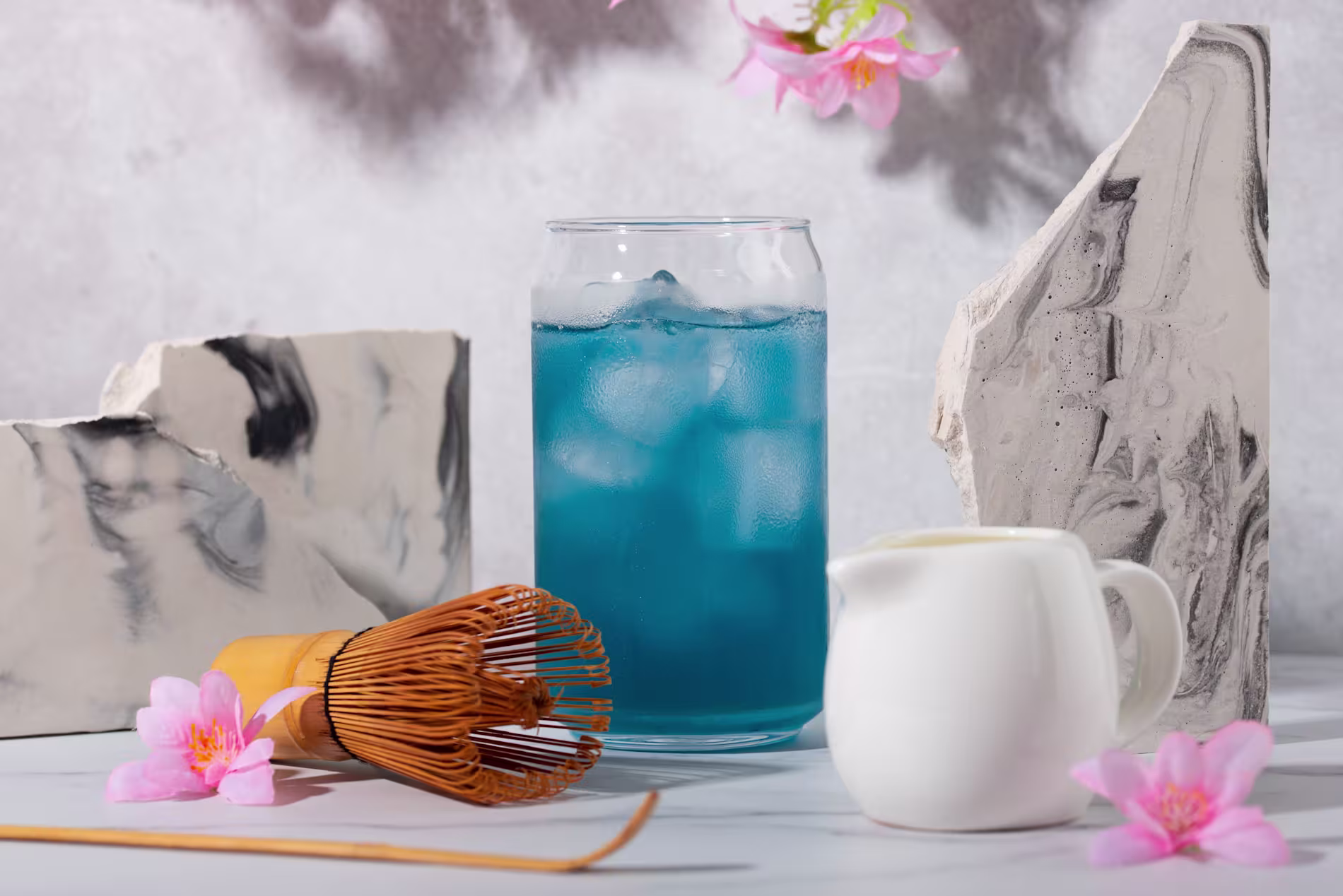 A glass of blue iced drink with a bamboo whisk, white pitcher, marble slabs, and pink flowers on a white surface.