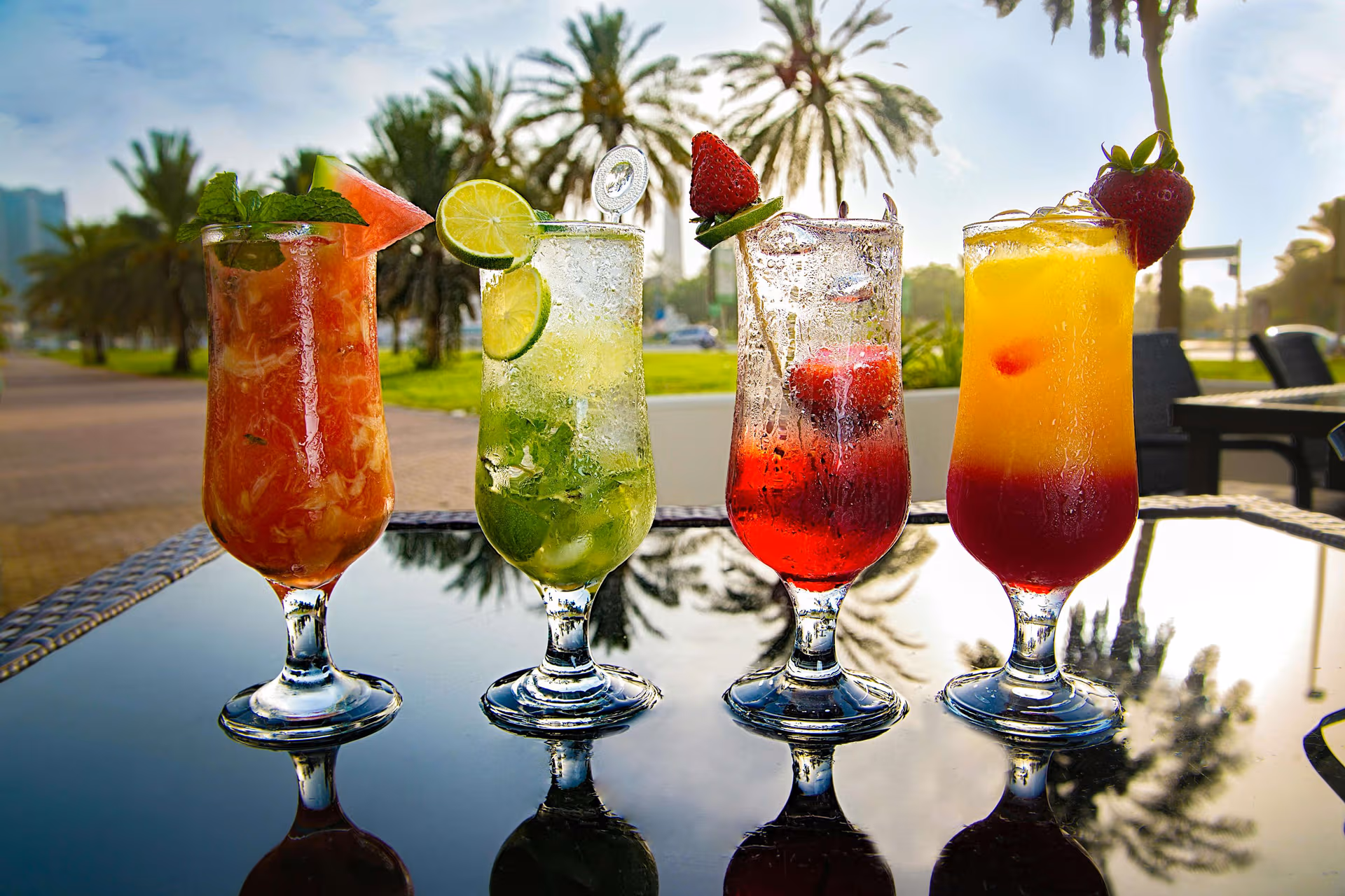 4 Vibrant Cocktails outside on a glass table surrounded by Palm trees 