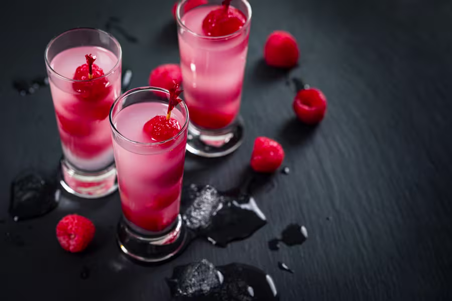Three glasses of pink raspberry cocktails, each garnished with whole raspberries, on a dark surface with scattered raspberries and water droplets.