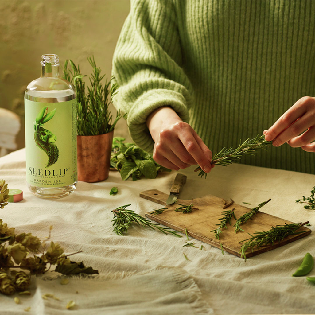 Seedlip Garden 108 bottle on a table with a person preparing fresh rosemary and herbs