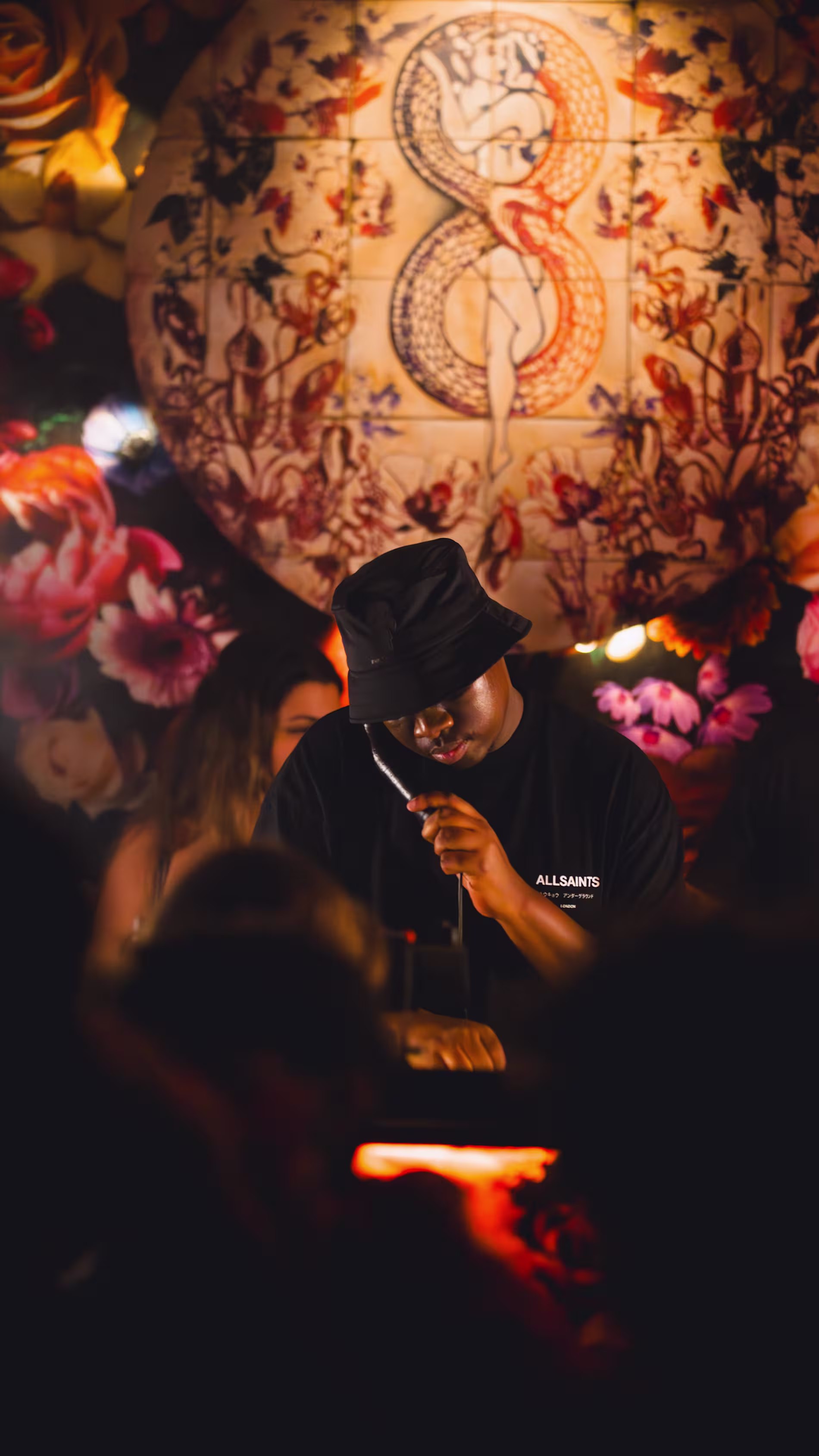 Un DJ concentrado frente al micrófono en Los Amantes, fondo lleno de arte floral y un diseño central intrigante que evoca romanticismo y misterio.