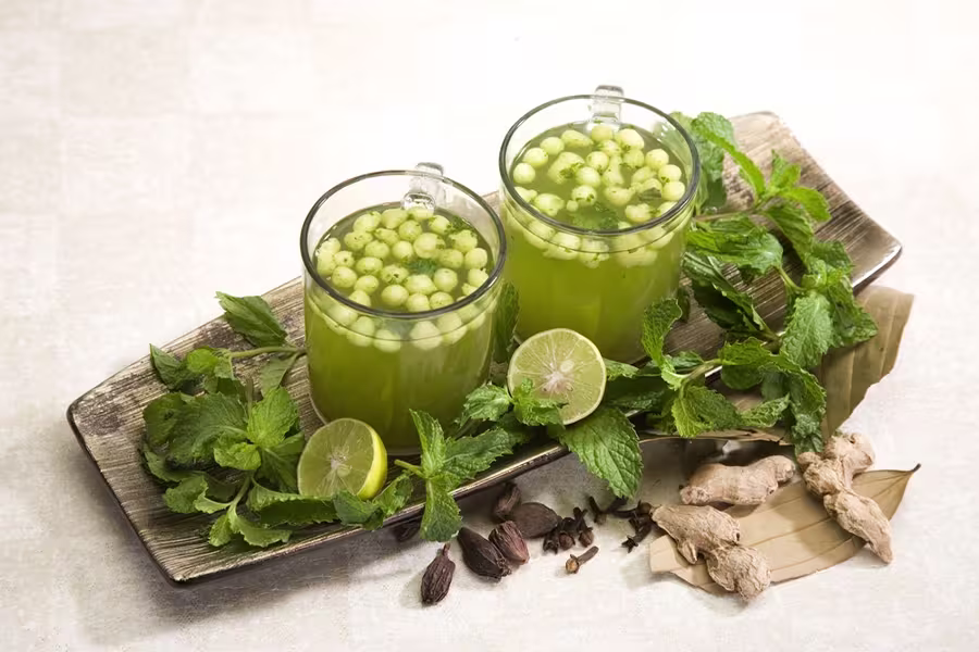 Two glasses of green drink garnished with mint leaves and round seeds, placed on a tray with fresh mint, lime halves, ginger, cardamom, cloves, and bay leaves.