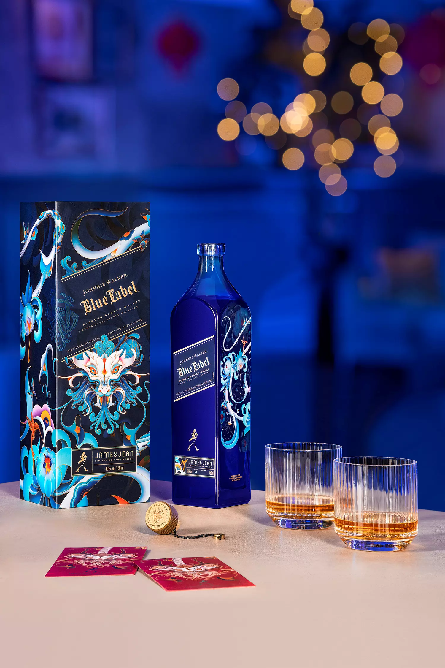 A blue Johnnie Walker Blue Label bottle with a decorative box stands next to two filled whiskey glasses and red envelopes on a table, with blurred lights in the background.