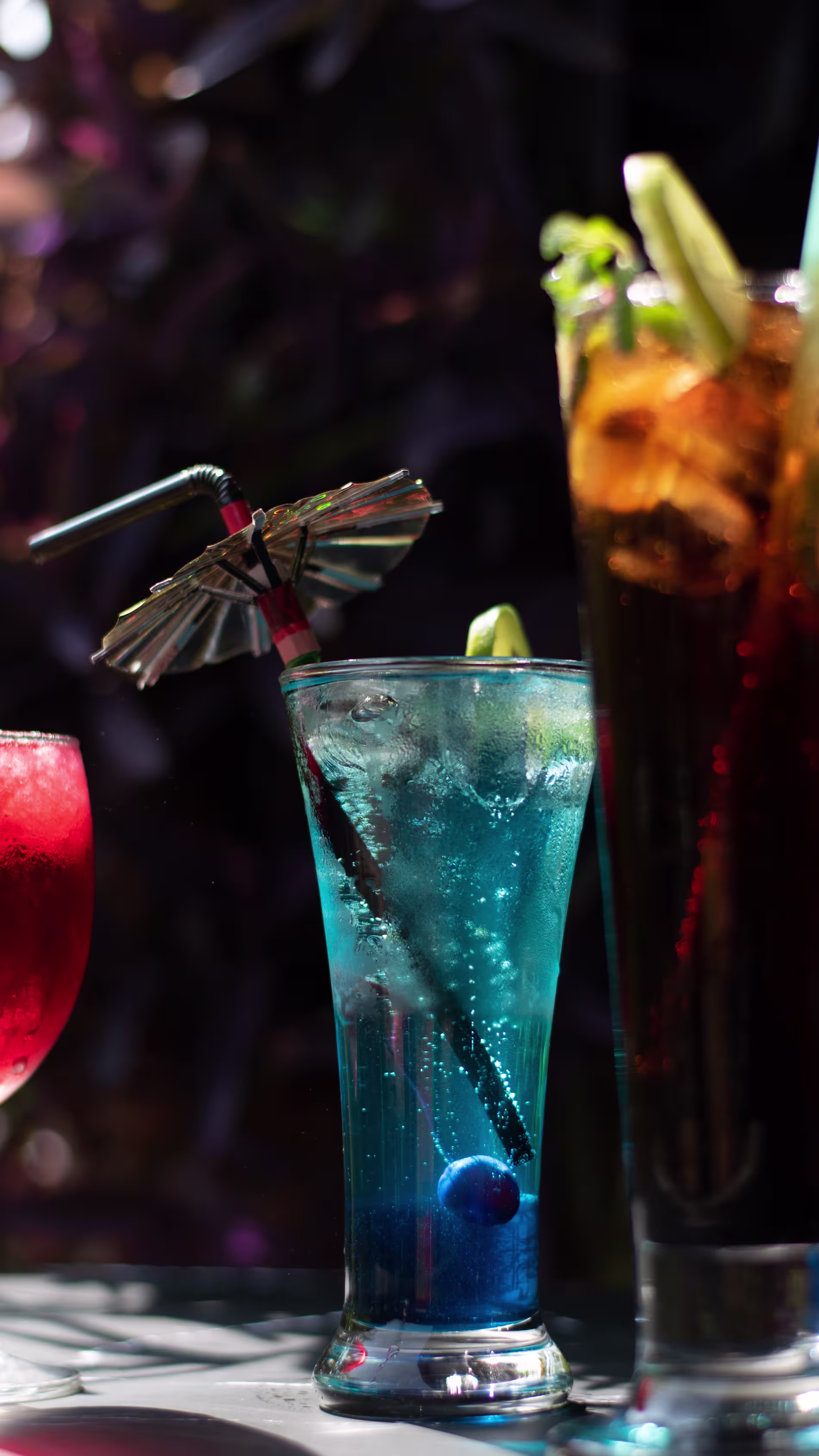 A blue cocktail with ice, a cherry, a lime wedge, a straw, and a paper umbrella is on a table next to two other colorful drinks.