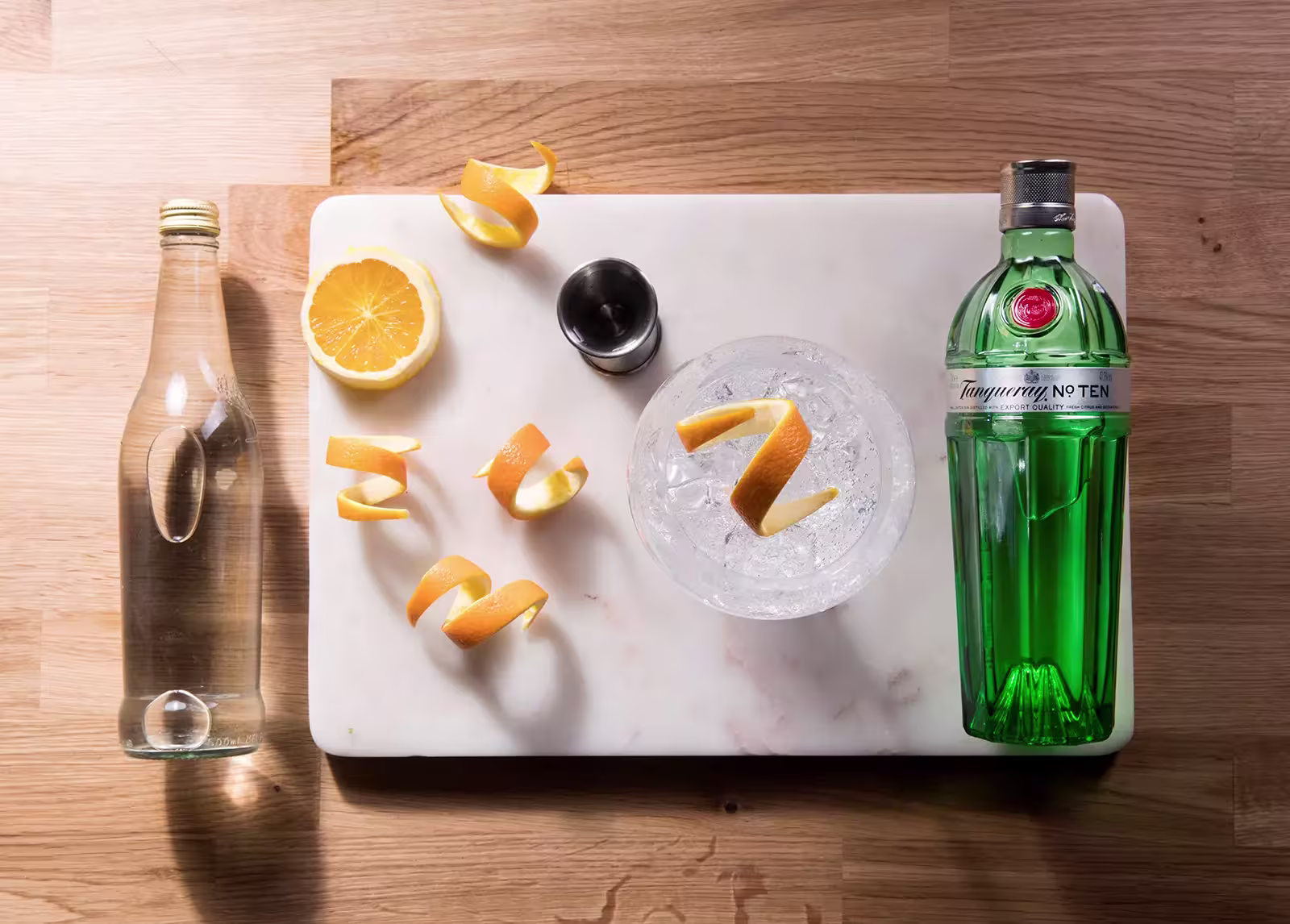 A bottle of gin, a water bottle, a halved orange, orange peels, a jigger, and a glass with ice and an orange twist are arranged on a white cutting board.