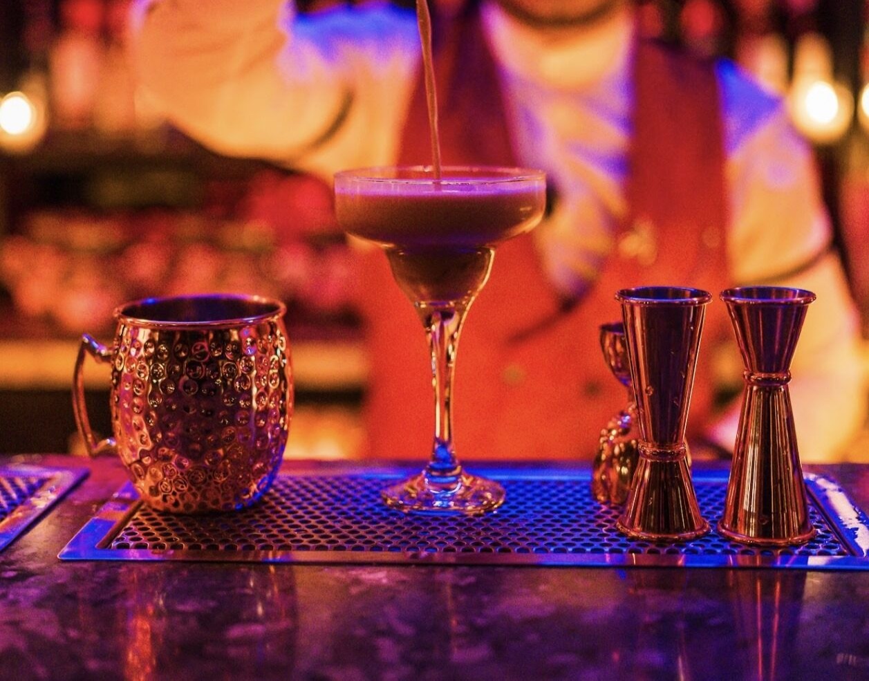 La imagen muestra a un bartender en Jimmy’s, sirviendo un cóctel con precisión y estilo en una copa elegante. El fondo iluminado en tonos cálidos y rojizos realza la atmósfera sofisticada del lugar.