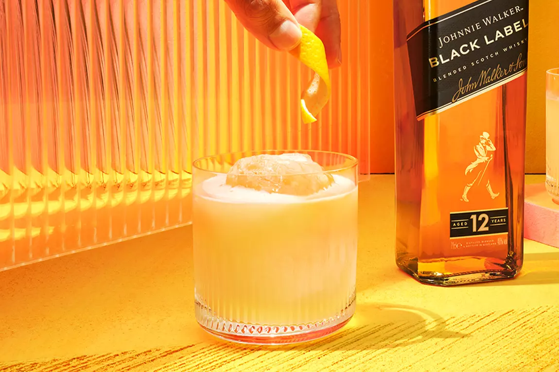 A hand garnishes a glass of whisky cocktail with an orange peel next to a bottle of Johnnie Walker Black Label on a yellow surface.