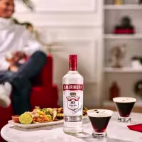 La imagen muestra una botella de vodka Smirnoff n°21 junto a dos vasos rellenos con el cóctel Espresso martini y un plato de comida a su lado.