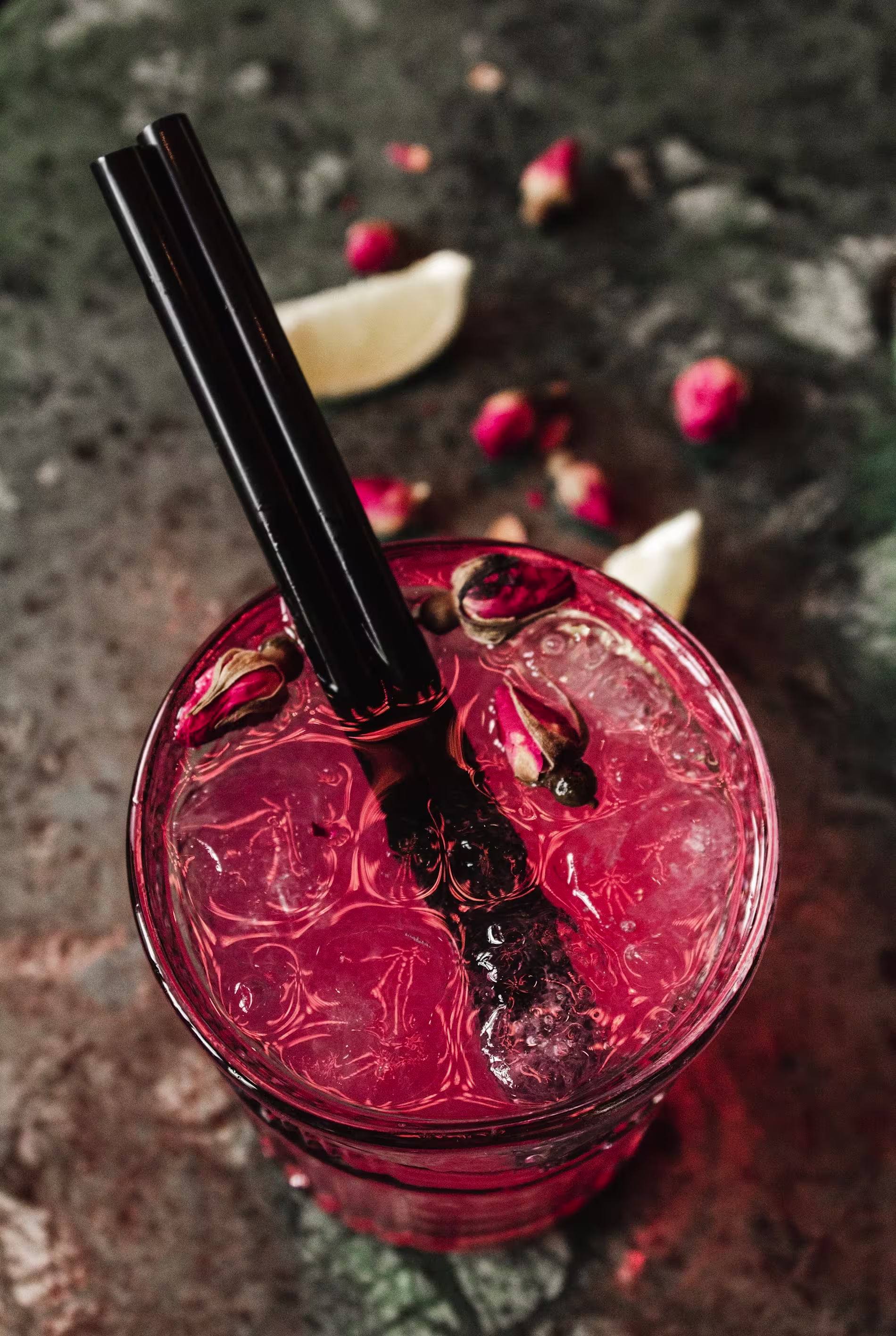 A pink iced drink with black straws, garnished with rosebuds, sits on a textured surface with lime wedges and rosebuds scattered nearby.